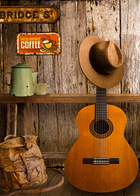 A guitar leans against a weathered bench and a well traveled  back pack sits next to it A fedora hangs on the head of the guitar. A green enamel coffee pot sits on the be